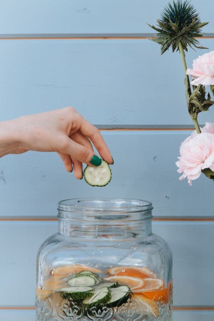 pexels-photo-10023980-10023980 Hands dropping cucumber slices into jar with citrus water, surrounded by flowers.